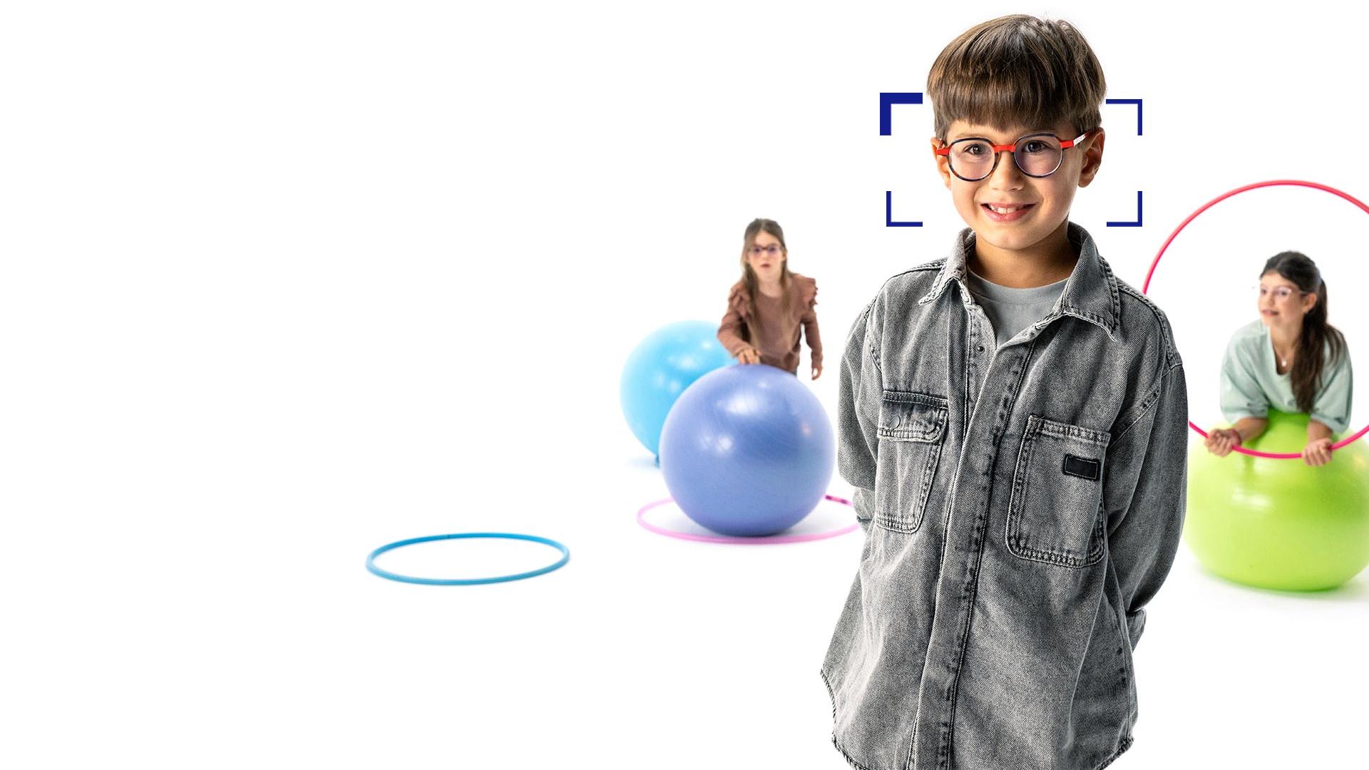 Brown-haired boy with round glasses wearing ZEISS MyoCare lenses stands in the foreground and smiles at the camera. In the background are two girls wearing ZEISS MyoCare lenses playing with hoops and gymnastic balls.